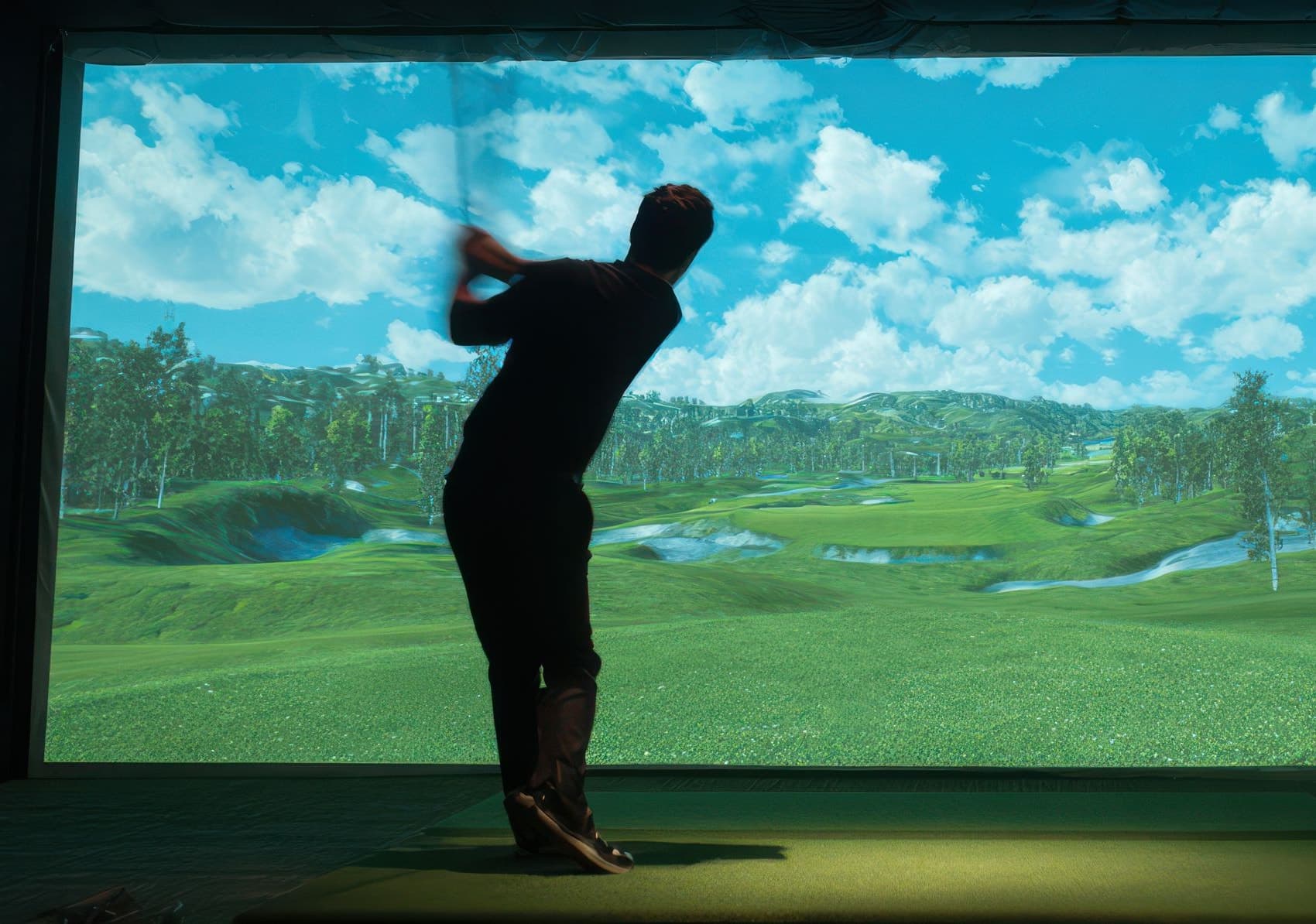 A player mid-swing in a darkened simulator bay, silhouetted against the lit course on screen